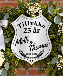 Nærbillede af personligt sølvbryllupsskjold blandt gran, hvide blomster og sølvbånd på en æresport.